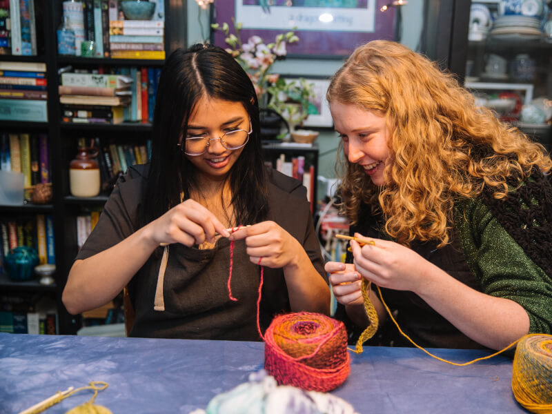Two women learn how to crochet while smiling.